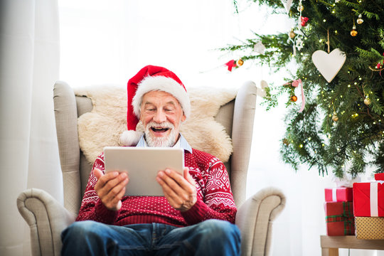 A portrait of senior man with tablet sitting on armchair at home at Christmas time.
