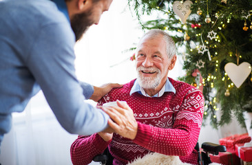 A hipster man giving drink to his senior father in wheelchair at Christmas time.