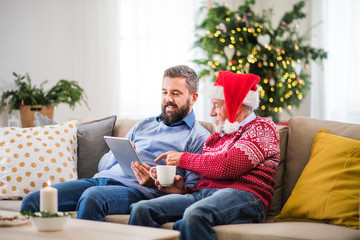 A senior father and adult son with tablet sitting on a sofa at home at Christmas time.