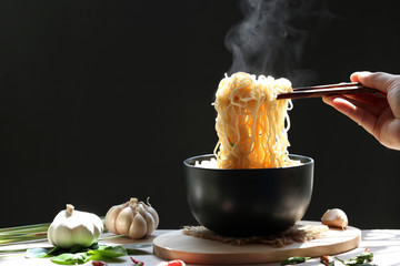 Woman hand holding chopsticks of instant noodles in cup  with smoke rising and garlic on dark...