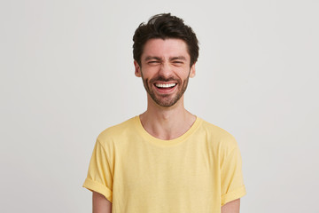 Close up of laughing happy screwing up his eyes attractive young man with dark brown hair and beard wears yellow tshirt, isolated over white background
