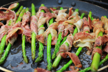 Raw bacon wrapped green asparagus in frying pan on dark background