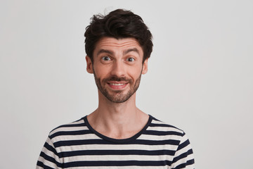 Close up of happy a little surprised attractive young brunet with beard eyebrows raised wears black and white striped tshirt, isolated over white background