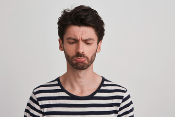 Studio shot of young brown hair man with beard looks sadly down, frowned eyebrows, lips pout, wears...