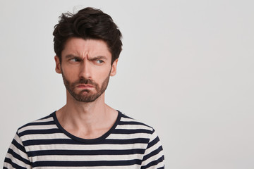 Obraz premium Young man with dark brown hair and beard wears black and white striped tshirt, looks angry, lips pursed frowning brows, head tuned rightside, stands leftside isolated over white background