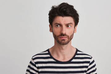 Young man with dark brown hair and beard wears black and white striped casual tshirt looks thinking, a little frowning brows, sight turned leftside, isolated over white background