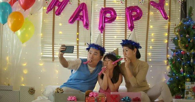 Best time together. Happy young family sitting on the bed and blowing party horn blowers while all of them wearing party hats in Slow motion.
