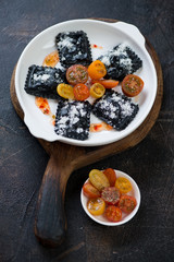 Plate of black ravioli with tomatoes and cheese topping on a rustic wooden serving board, studio shot over dark brown stone surface