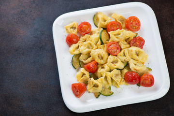 Tortellini with fried zucchini and tomatoes served in a white plate over dark brown metal background, horizontal shot with space