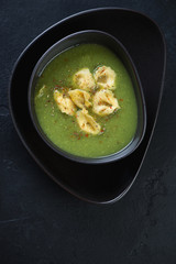 Broccoli cream-soup with tortellini served in a black bowl, flatlay over black stone background, vertical shot