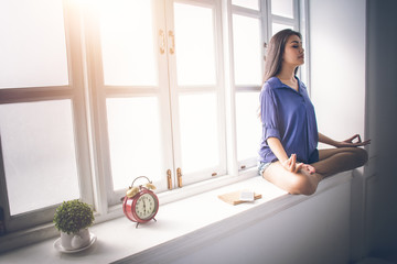 Asian girl sitting by the window. Her eyes closed in meditation and yoga postures. Amidst the mild...