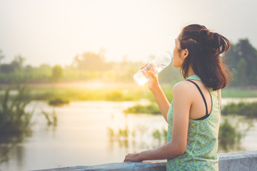 Asian girls are drinking mineral water after exercise very tiring. She stood on a concrete bridge which is across the river. The atmosphere of the evening sun and green trees at the waterfront.