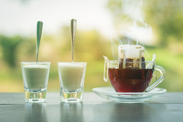 A clear glass cup on drip coffee, placed on a wooden table. There is white smoke floating above the cups of coffee creamer and sugar in a glass, which is placed beside the Sun morning, warm atmosphere