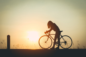 Obraz premium The silhouette of a beautiful girl with long hair asian were towed bicycle on a road amid the ambiance of the evening sunset, flowers grass at the side of the road, Her face fell with the feeling sad.