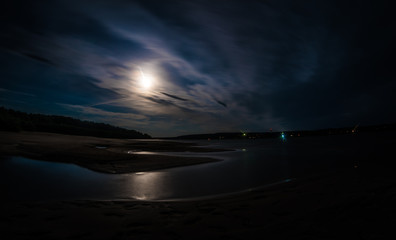 A long exposure shot of Tom river with moonlit water and shores under the cloudy night sky,...