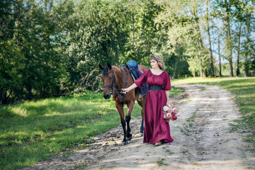 A woman with a bouquet of flowers dressed in a long burgundy dress with sleeves leads a brown horse along a rural road