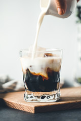 Pouring milk in a glass with espresso and cubes of ice in a modern kitchen in the morning.
