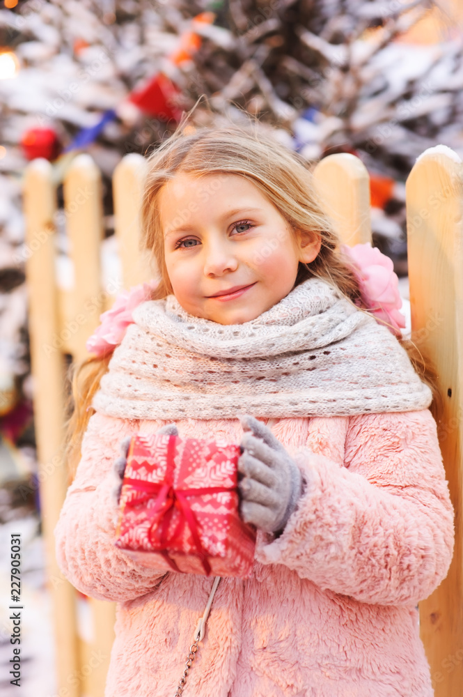 Wall mural happy child girl holding christmas gift outdoor on the walk in snowy winter city decorated for new year holidays.