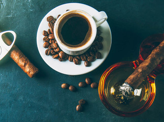 Cup of coffee, coffee beans, ashtray with cigar on dark background.