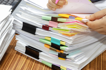 Teacher's hand is checking student homework assignment and report on desk in office for score. Unfinished paperwork stacked in archive with color papers and paper clips. Education and business concept