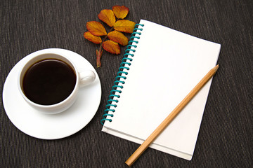 white Cup of black coffee stands on a dark background with colored autumn leaves and a notebook