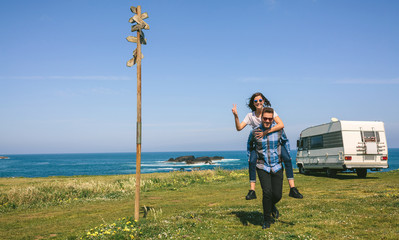 Young man piggybacking his girlfriend near the coast with camper van in the background