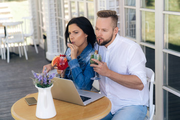 Thoughtful business people are sitting at a table in a cafe, watching a boring film on a laptop, because of which both are immersed in their thoughts, they are drinking tasty, cool cocktails.