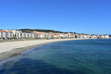 Fototapeta na wymiar Small coastal village with beach and promenade. Turquoise and clear water, bright sand, blue sky. Sanxenxo, Rias Baixas, Galicia, Spain.