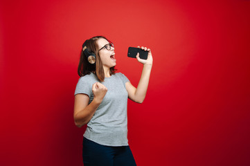 Obraz premium Portrait of emotional young woman holding her smartphone like microphone and singing, on red background