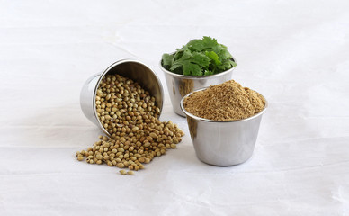 Coriander powder, plucked coriander leaves, and spilled coriander seeds, in steel bowls on a crumpled paper background.