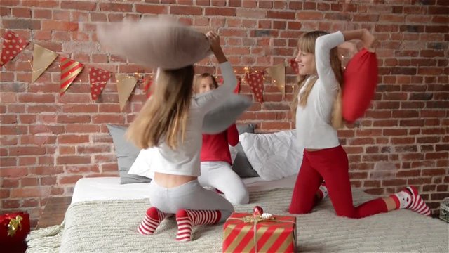 Happy laughing Triplets sisters in pajamas having a pillow fight in bed at home. The bedroom is decorated by christmas garlands and xmas gifts, Merry Christmas