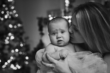 Happy young mother kissing baby daughter for the Christmas tree lights in the background. Family, motherhood and holidays concept. Black and white photo.