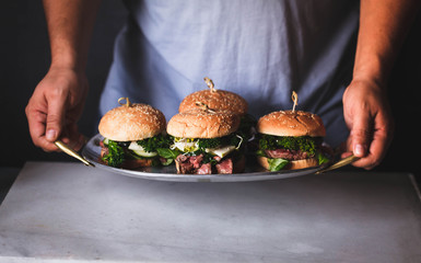 Male hands holding four burgers with grilled beef meat on tray on marble cutting board 