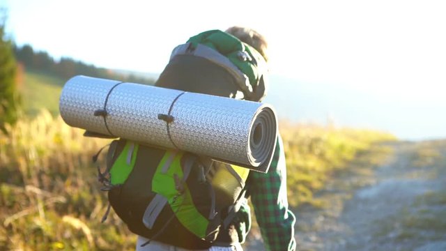Slow motion footage with natural optical flares  of a little hiker boy going by the mountain road with backpack with rolled sleeping pad