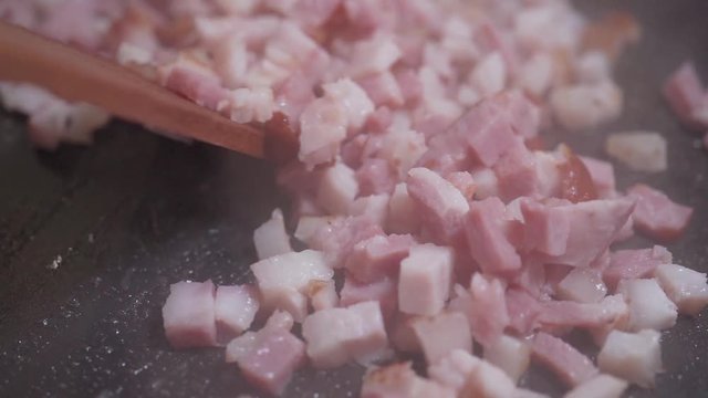 A cook with a wooden spatula stirs the chopped bacon are fried in a hot frying pan