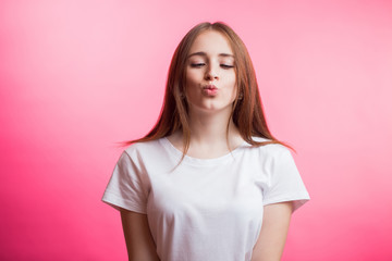 Obraz premium Cute lovely young woman in white shirt standing and sending kiss over pink background. Girl looking down