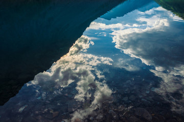 Stones and plants on bottom of mountain lake with clean water in sunny day close up. Giant mountains and cloudy sky reflected on smooth water surface in sunlight. Background with underwater vegetation