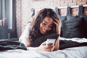 Obraz premium Portrait of beautiful woman waking up in her bed and looks into the phone. Check social networks, send sms. The girl is wearing a T-shirt