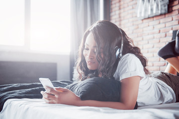 Beautiful african girl in sleepwear smiling listening to music in headphones lying on bed