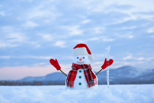 Unbelievable sunrise. Happy snowman in hat, scarf, red gloves with ice pikestaff is standing on the snow lawn. Mountains landscape on the background.