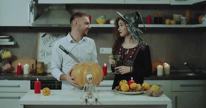 A couple prepare for Halloween party at their kitchen, they are decorating the kitchen tabel, starting to cut a very big pumpkin , young lady is masked for Halloween. 4k