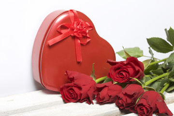 A bouquet of scarlet roses and a gift in a box in the shape of a heart. Five flowers lie on a wooden box. On a white background.