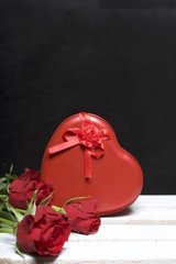 A bouquet of scarlet roses and a gift in a box in the shape of a heart. Flowers lie on a wooden box. On a black background.