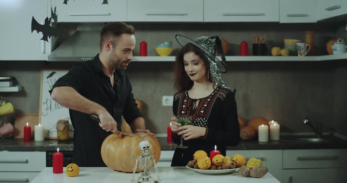Smiling young couple preparing for a Halloween party at the kitchen tabel trying to cut a big Halloween pumpkin, spending a great time , the lady are masked already holding a green glass of cocktail