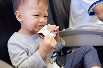 Cute little Asian 2 years old toddler baby boy child bite / eating chocolate bar during flight on airplane. Flying with children, Happy air travel with kids & little traveler concept