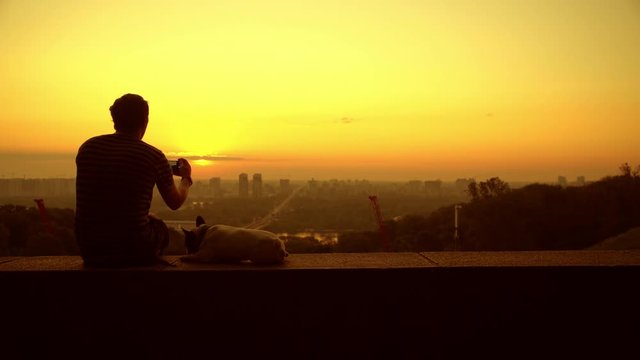 A man photographs the beautiful landscape that he watchs with his dog. The man wears a striped T-shirt, it's the sunrise.