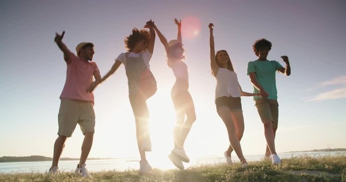 Group of five multiethnic friends running down meadow near lake, jumping, cheering, and smiling. Friends having fun 4k