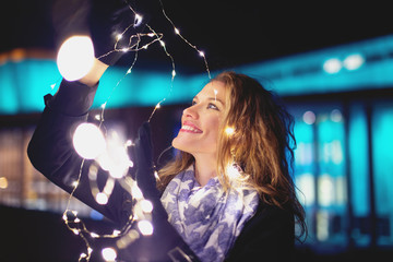Happy young woman playing with fairy lights at winter evening in city