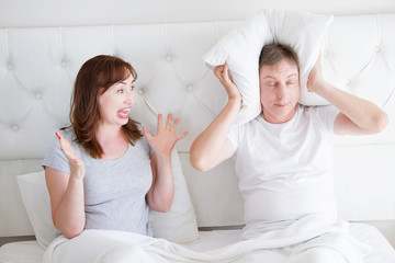 Caucasian middle age family couple angry shouting in bed. Conflict relationship concept. Husband cover ears by hands and wife trying make a dialogue. Selective focus