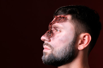 Profile of a young man with ragged facial wounds. A guy with Halloween make-up looks to the left against a burgundy background. Close-up. Copyspace.